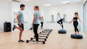 A group of people performing various coordination exercises in a well-lit, modern sports therapy clinic. In the foreground, two individuals stand facing each other, one balancing on one leg while the other guides their movements. In the middle ground, a person navigates a series of agility ladders, their feet stepping with precision. In the background, a person balances on a BOSU ball, their core engaged as they shift their weight side to side. The atmosphere is one of focused concentration and therapeutic rehabilitation, with clean lines, neutral colors, and a sense of movement and progression.